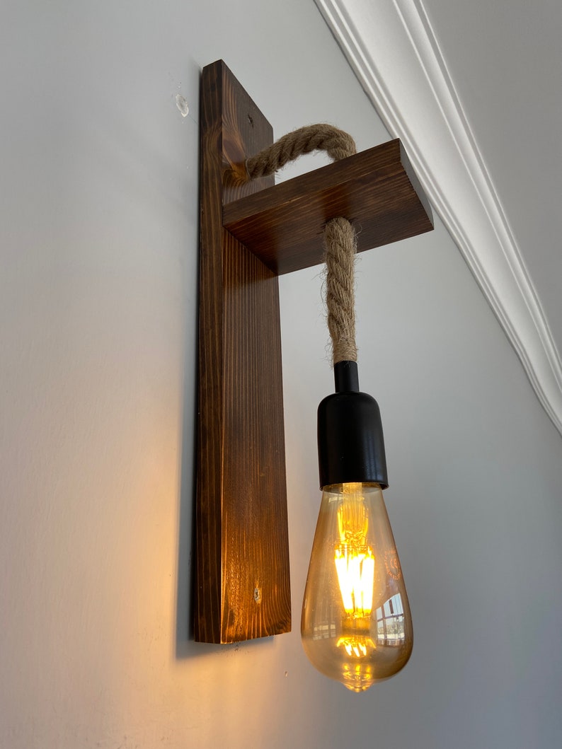 May include: A wall-mounted light fixture featuring a dark stained wooden backplate and a horizontal wooden arm. A thick, natural-colored jute rope hangs from the arm, supporting a black socket with a glowing, teardrop-shaped Edison light bulb.