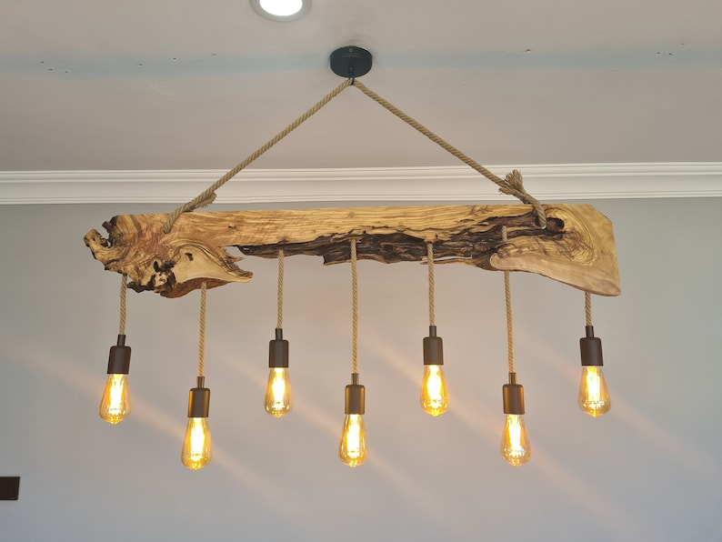 May include: A rustic wooden chandelier with eight Edison-style light bulbs. The light fixture is suspended from the ceiling by a thick rope and a black metal canopy. The wooden beam has a natural, unfinished look.