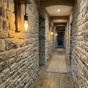 May include: A wall-mounted light fixture featuring a rectangular wooden backplate, a horizontal wooden shelf, and a thick rope cord suspending a black socket with a glowing Edison-style light bulb, installed on a textured stone wall.