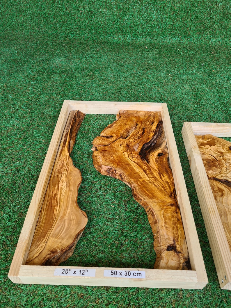 May include: A rectangular wooden frame with a natural wood finish encloses a piece of olive wood with a unique grain pattern. The wood is a light brown color with darker brown streaks. The frame is labeled with the dimensions 20" x 12" and 50 x 30 cm.