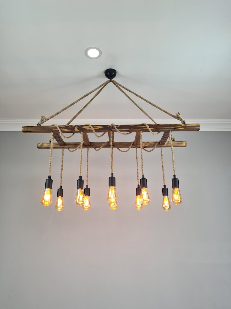 K&ouml;nnte beinhalten: Ein rustikaler Holzleiter-Kronleuchter mit 10 warmwei&szlig;en Edison-Gl&uuml;hbirnen, die an Seilschn&uuml;ren h&auml;ngen. Der Kronleuchter ist mit einem schwarzen Metallhaken an der Decke befestigt.