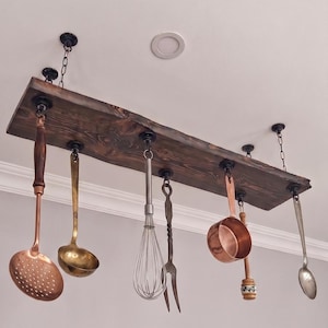 May include: A rectangular dark stained wooden board suspended from the ceiling by black chains and hardware. Six kitchen utensils hang from the board, including a copper skimmer, a brass ladle, a wire whisk, a metal fork, a copper pot, and a spoon.