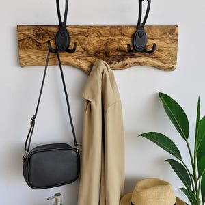 May include: A wooden wall-mounted coat rack with two black metal hooks. A black crossbody bag and a tan jacket hang from the hooks. A straw hat with a black band sits on a wooden drawer. A white soap dispenser and a wooden dish are on the table.