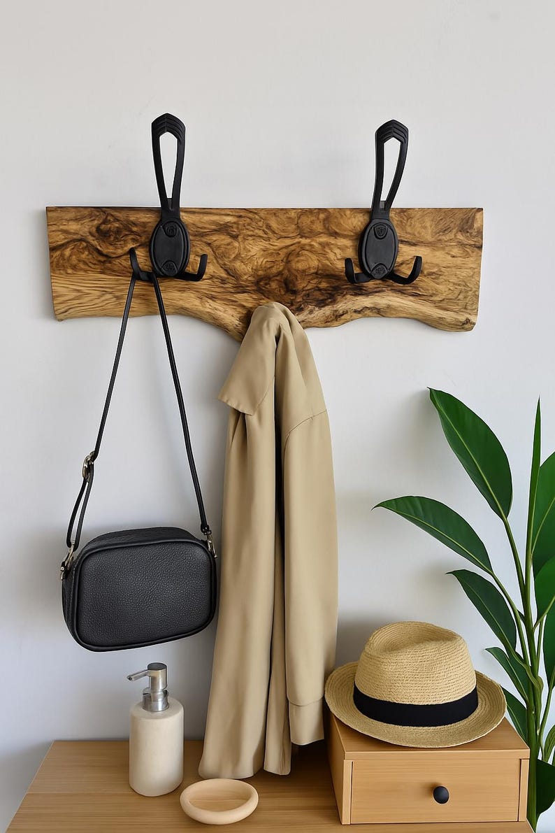 May include: A wooden wall-mounted coat rack with two black metal hooks. A black crossbody bag and a tan jacket hang from the hooks. A straw hat with a black band sits on a wooden drawer. A white soap dispenser and a wooden dish are on the table.