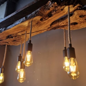 Może przedstawiać: Lampa sufitowa z naturalną drewnianą płytą o nieregularnych krawędziach i ciemnym, teksturowanym wykończeniu kory. Sześć żar&oacute;wek w stylu Edisona zwisa z drewna na grubych, beżowych sznurach, zamocowanych w czarnych, cylindrycznych oprawkach.