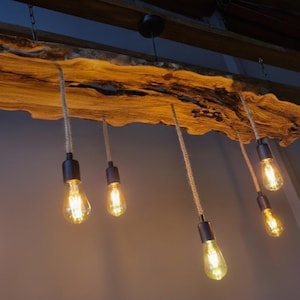 Może przedstawiać: Lampa sufitowa wykonana z drewnianej belki z naturalną krawędzią, z kt&oacute;rej zwisa siedem żar&oacute;wek w stylu Edisona na grubych sznurach. Widoczne jest naturalne usłojenie drewna, a żar&oacute;wki emitują ciepłe światło z czarnych metalowych oprawek.