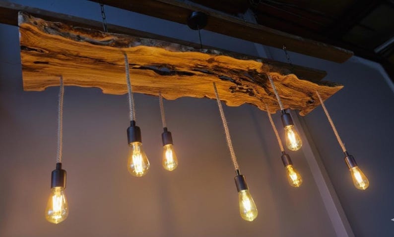 Może przedstawiać: Lampa sufitowa wykonana z drewnianej belki z naturalną krawędzią, z kt&oacute;rej zwisa siedem żar&oacute;wek w stylu Edisona na grubych sznurach. Widoczne jest naturalne usłojenie drewna, a żar&oacute;wki emitują ciepłe światło z czarnych metalowych oprawek.