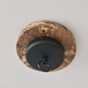 Puede incluir: Un accesorio de techo de metal negro con un gancho para colgar una lámpara. El accesorio está montado en una base redonda de madera con veta de madera natural.