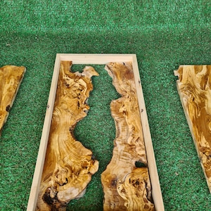 Puede incluir: Un marco de madera rectangular con un acabado de madera natural encierra una pieza de madera de olivo con un patr&oacute;n de veta &uacute;nico. La madera tiene un color marr&oacute;n natural con una veta marr&oacute;n claro y beige. El marco est&aacute; etiquetado con las medidas 24" x 12" y 50 x 30 cm.