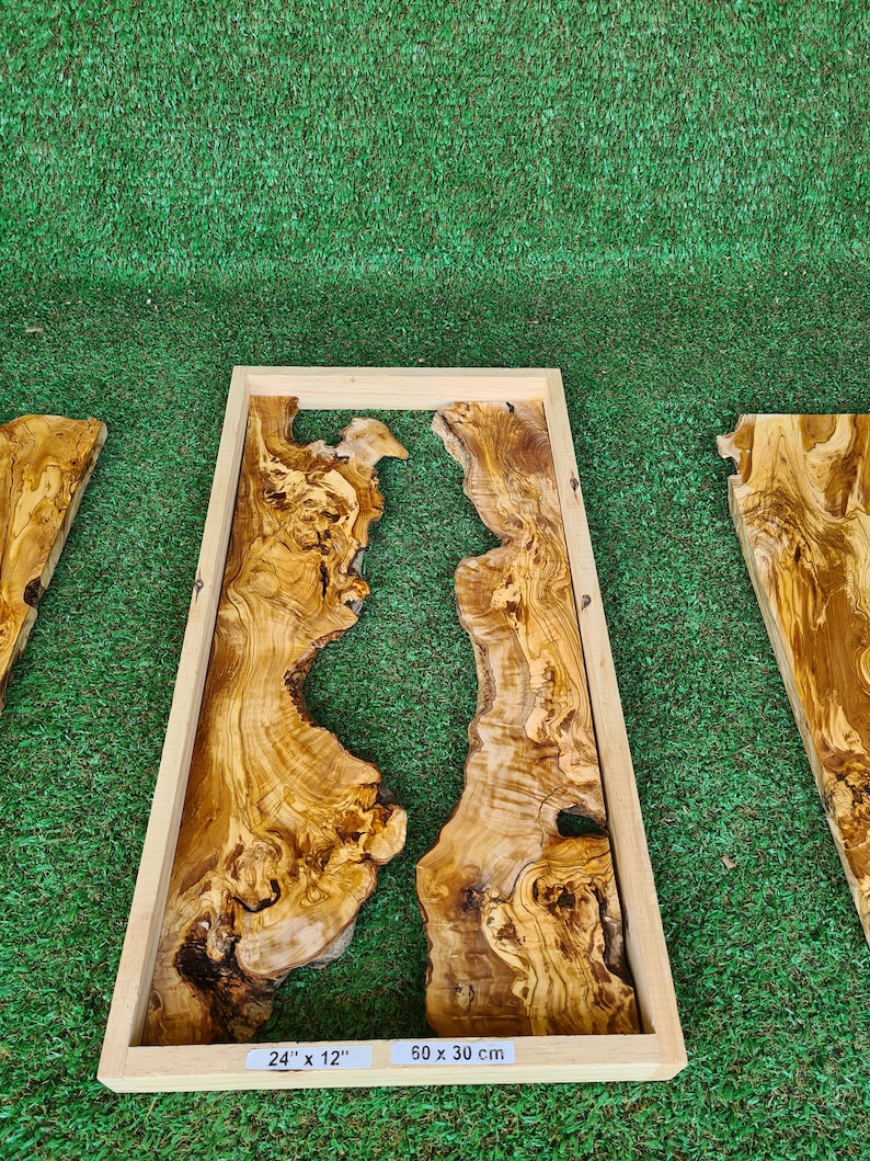 Puede incluir: Un marco de madera rectangular con un acabado de madera natural encierra una pieza de madera de olivo con un patr&oacute;n de veta &uacute;nico. La madera tiene un color marr&oacute;n natural con una veta marr&oacute;n claro y beige. El marco est&aacute; etiquetado con las medidas 24" x 12" y 50 x 30 cm.