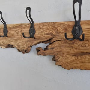 May include: A rustic wooden coat rack with four black metal hooks. The wood has a natural, irregular edge and a warm, brown tone. The hooks are mounted on the wood, creating a functional and decorative piece for entryways or bedrooms.