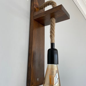 May include: A wall-mounted light fixture featuring a dark brown stained wooden backplate and a perpendicular wooden shelf. A thick, natural-colored jute rope hangs from the shelf, supporting a black socket with a clear, teardrop-shaped Edison bulb.