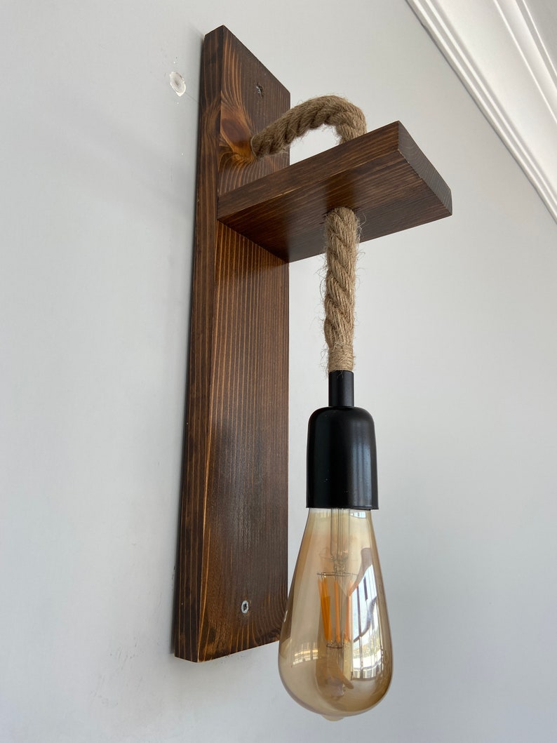 May include: A wall-mounted light fixture featuring a dark brown stained wooden backplate and a perpendicular wooden shelf. A thick, natural-colored jute rope hangs from the shelf, supporting a black socket with a clear, teardrop-shaped Edison bulb.