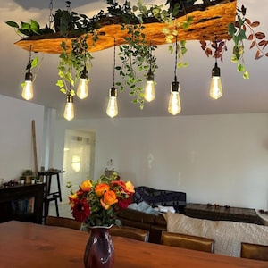 May include: A rustic wooden light fixture with six hanging Edison bulbs and greenery accents. The fixture is suspended from the ceiling and illuminates a dining room table with a vase of flowers.