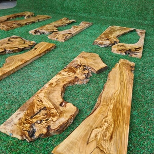 Puede incluir: Varias piezas de tablas de madera de olivo con veta de madera natural y nudos. Las tablas est&aacute;n dispuestas sobre una superficie de c&eacute;sped artificial verde.