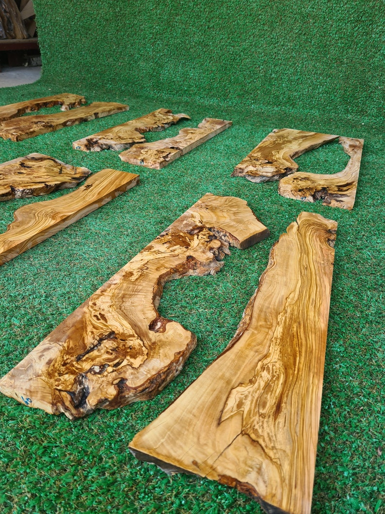 Puede incluir: Varias piezas de tablas de madera de olivo con veta de madera natural y nudos. Las tablas est&aacute;n dispuestas sobre una superficie de c&eacute;sped artificial verde.