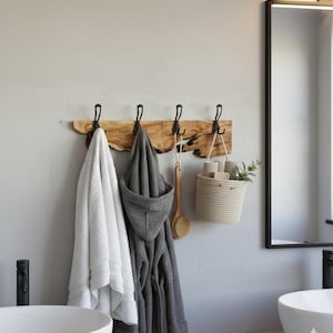 May include: A rustic wooden wall-mounted rack with black metal hooks, holding a white towel, a gray hooded robe, and a wooden brush. A woven basket with toiletries and greenery hangs nearby, with a large mirror in the background.