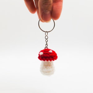 May include: A crocheted mushroom keychain with a red cap and white spots, and a white stem. The keychain is attached to a silver key ring.