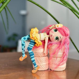 Op de afbeelding: Een handgemaakte gehaakte pop met roze haar, groene ogen en een gele en oranje jurk. De benen van de pop zijn blauw met stippen en strepen en ze draagt oranje schoenen. De pop leunt op een stapel roze garenklosjes.