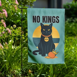 May include: A teal garden flag with the words "NO KINGS" at the top. The flag features a black cat with a gold collar and the word "RESIST" on it, sitting in front of a yellow circle and a brown and blue stripe. A small orange crown is at the cat's feet.