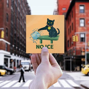 May include: A square, orange sticker held in front of a city street. The sticker features a black cat with a yellow collar, pouring ice from a blue ice cube tray. The word "NO ICE" is printed below the cat.