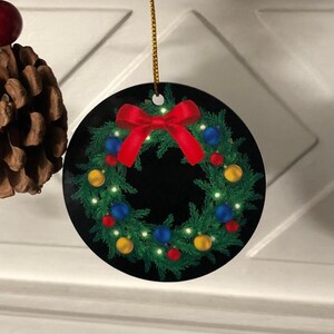 May include: A round, flat Christmas ornament featuring a printed image of a green pine wreath against a black background. The wreath is decorated with small red, blue, and yellow spherical ornaments, tiny white lights, and a large red bow at the top.