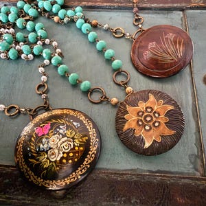 May include: A turquoise and white beaded necklace with three round pendants. The pendants are painted with floral designs and have a dark brown background.