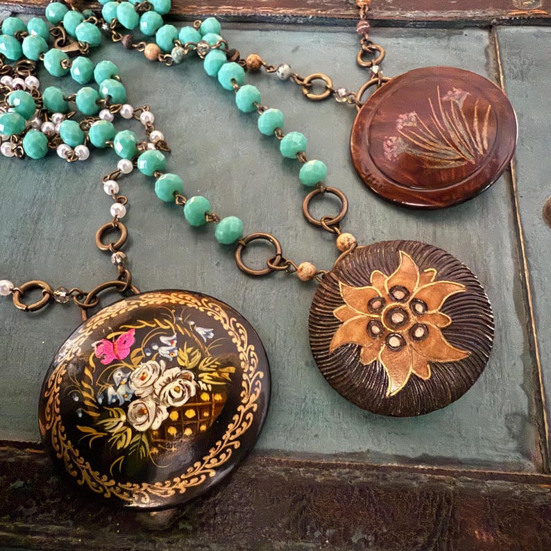 May include: A turquoise and white beaded necklace with three round pendants. The pendants are painted with floral designs and have a dark brown background.
