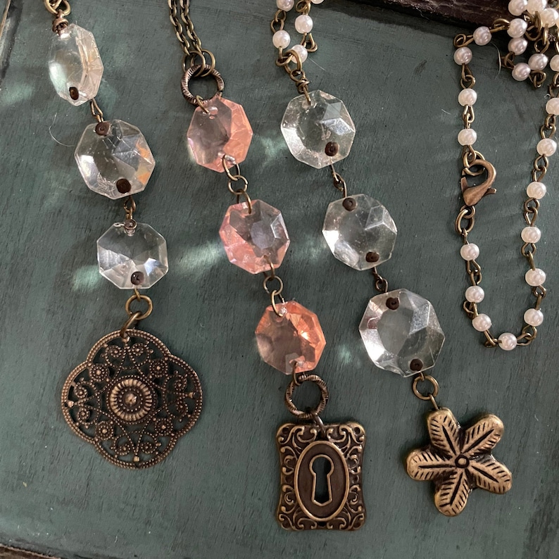 Puede incluir: Tres collares con abalorios de cristal y metal. Los collares presentan cuentas de cristal transparentes y rosadas, un abalorio de ojo de cerradura, un abalorio de flor y un abalorio de filigrana.