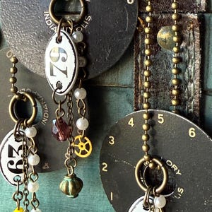 Könnte beinhalten: Nahaufnahme von handgefertigten Ohrringen im Steampunk-Stil. Jeder Ohrring hat ein weißes ovales Etikett mit einer Nummer, verziert mit Perlen, Zahnrädern und Charms. Die Ohrringe hängen vor einem dunklen, strukturierten Hintergrund mit Zifferblättern.