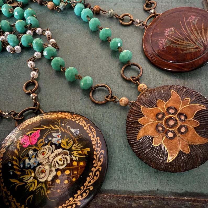 May include: Three necklaces with round pendants. The pendants are painted with floral designs. One pendant is black with gold accents and features a butterfly and flowers. Another pendant is brown with a gold flower design. The third pendant is brown with a floral design in pink and white.