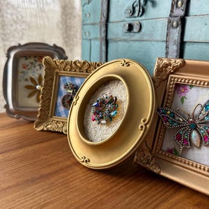 May include: A collection of ornate, vintage-style frames in gold and silver tones. One frame features a colorful jeweled brooch, while others display floral and patterned designs. A teal trunk is in the background.