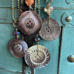 May include: A necklace with four brown and beige button pendants, each with a different design. The buttons are strung on gold chains with crystal beads.