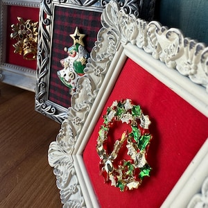 May include: Ornate framed Christmas brooches. The brooches include a gold crown, a Christmas tree with a star, and a wreath with a candle. The frames are silver and white with red and plaid fabric backgrounds.