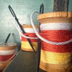 May include: Close-up of three wooden spools of thread, each with a candy corn color scheme: white, orange, and yellow. Black needles are inserted into the spools, with white thread looped around them. The spools are arranged on a dark green surface.