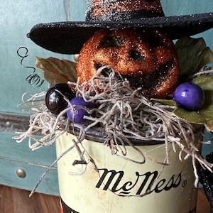 May include: A Halloween decoration featuring a pumpkin with a black witch hat, surrounded by purple and black marbles, nestled in a vintage tin can with the text "McNess" printed on it.