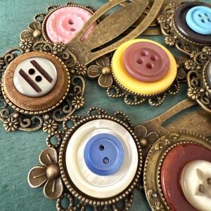 May include: Close-up of several antique bronze hair clips, each featuring a decorative filigree frame and a colorful button center. Buttons are in shades of pink, brown, yellow, blue, and white. The clips are arranged on a teal surface.
