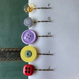 K&ouml;nnte beinhalten: F&uuml;nf verschiedene Haarklammern mit verschiedenen Knopf-artigen Verzierungen. Die Klammern sind goldfarben und die Kn&ouml;pfe sind in verschiedenen Farben und Mustern, darunter gelb, lila und rot.