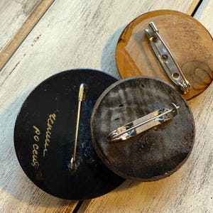 May include: Three round wooden pins with metal clasps on the back. The pins are black, brown, and light brown. The black pin has gold lettering that reads "100 ceus" and "threes".