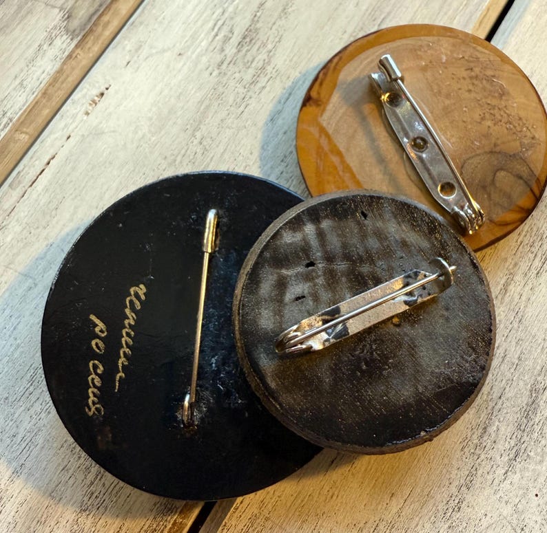 May include: Three round wooden pins with metal clasps on the back. The pins are black, brown, and light brown. The black pin has gold lettering that reads "100 ceus" and "threes".