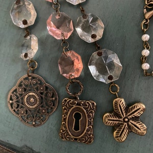 Puede incluir: Un collar delicado con una mezcla de cuentas de vidrio transparentes y rosadas, con tres dijes: una cerradura, una flor y un dise&ntilde;o de filigrana. El collar est&aacute; hecho de lat&oacute;n y tiene un estilo vintage.