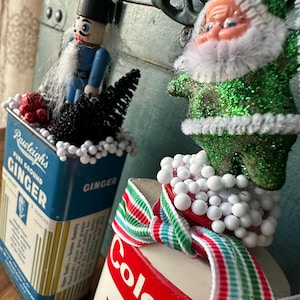 May include: Two vintage Christmas decorations made from repurposed tin cans. The first can is blue and has the text "Rawleigh's Pure Ground Ginger" on the front. The second can is white with red and green stripes and the text "Colgate" on the front. The can on the left is topped with a nutcracker figurine, a small black tree, and white styrofoam balls. The can on the right is topped with a green glitter Santa Claus figurine and white styrofoam balls.