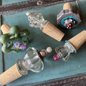 May include: Five decorative bottle stoppers with cork bases and various glass and metal embellishments. The stoppers feature a variety of colors, including green, blue, red, and white.