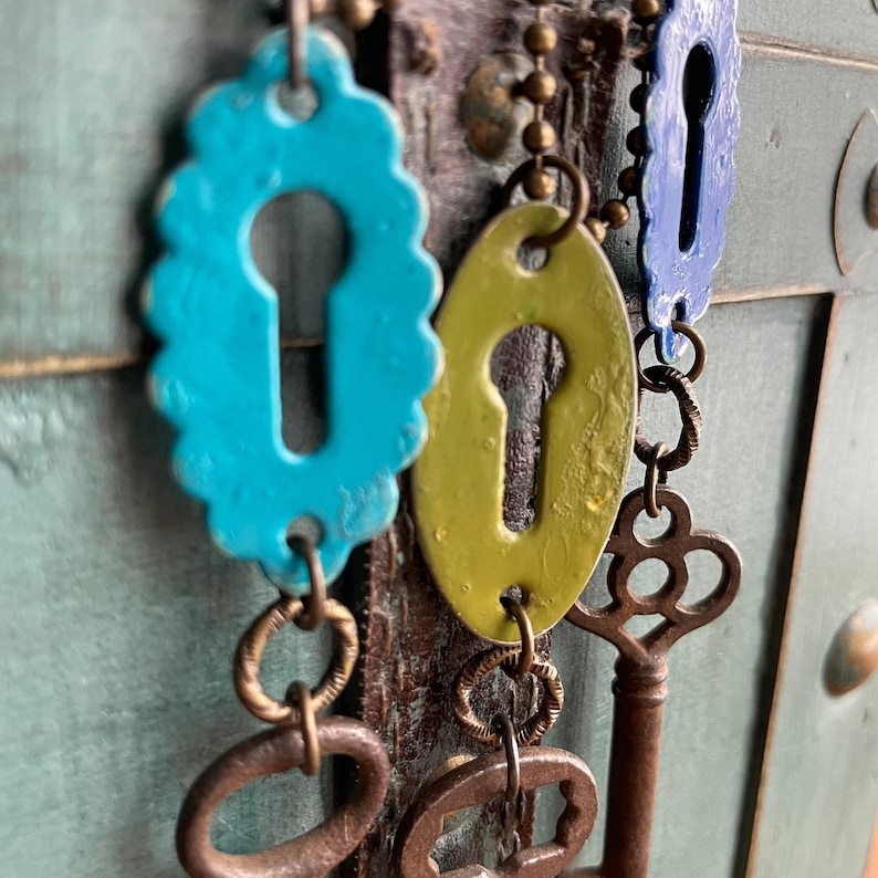 May include: A collection of keyhole-shaped charms in blue, green, and brown, hanging from a chain with antique keys. The charms are attached to a wooden surface.