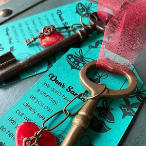May include: Two antique-style keys with red heart charms and red ribbon, resting on teal cards with text. The cards read "Dear Santa" and "Magic Key". The keys are likely decorative or symbolic.