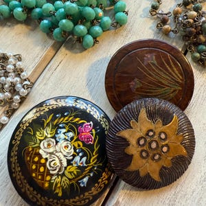 May include: Three round wooden decorative ornaments with painted designs. One ornament features a floral design with pink, white, and yellow flowers. Another ornament features a brown flower with a yellow center. The third ornament features a black background with a painted floral design in shades of yellow, white, and blue.