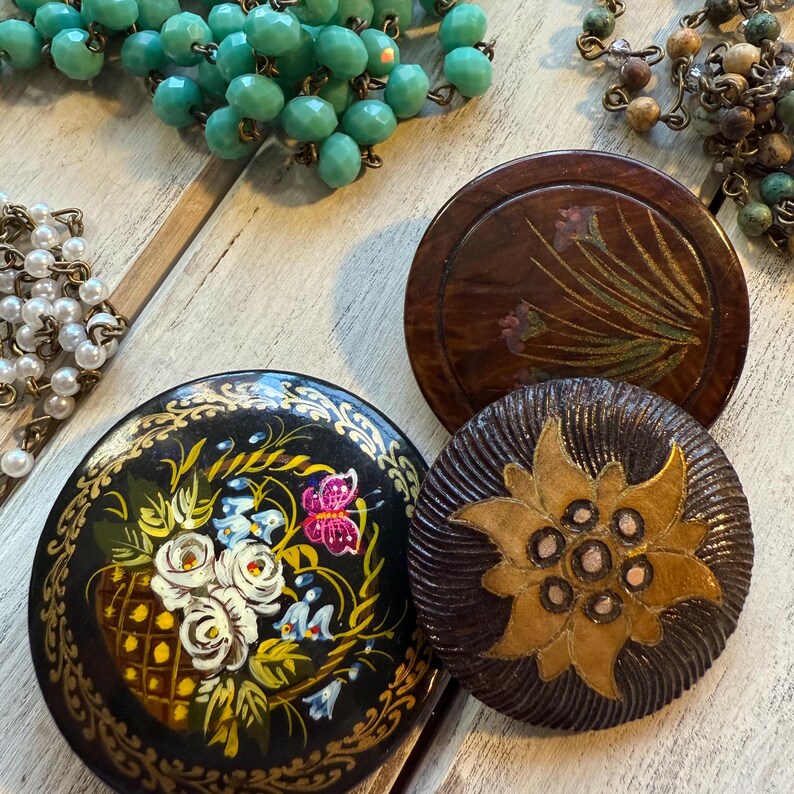 May include: Three round wooden decorative ornaments with painted designs. One ornament features a floral design with pink, white, and yellow flowers. Another ornament features a brown flower with a yellow center. The third ornament features a black background with a painted floral design in shades of yellow, white, and blue.