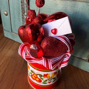 May include: A red and white striped ribbon surrounds a red tin can with a floral design. The can is filled with red hearts, glitter hearts, and red ornaments.