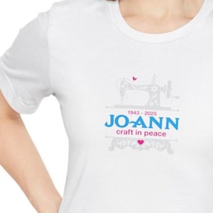 Puede incluir: Camiseta blanca con un diseño de máquina de coser y el texto azul "JO-ANN", los años "1943-2025" y la frase "craft in peace". También hay un pequeño corazón rosa.