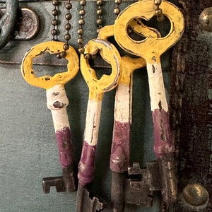 May include: Five antique keys with yellow and white painted details. The keys are attached to a chain and hang from a metal surface.